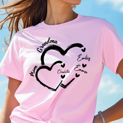 May include: Pink t-shirt with a black design of two interlocking hearts. The text "Grandma" is above the hearts, and the text "Mom" is to the left of the hearts. Inside the hearts are the names "Emily", "Emma", and "Charlotte".