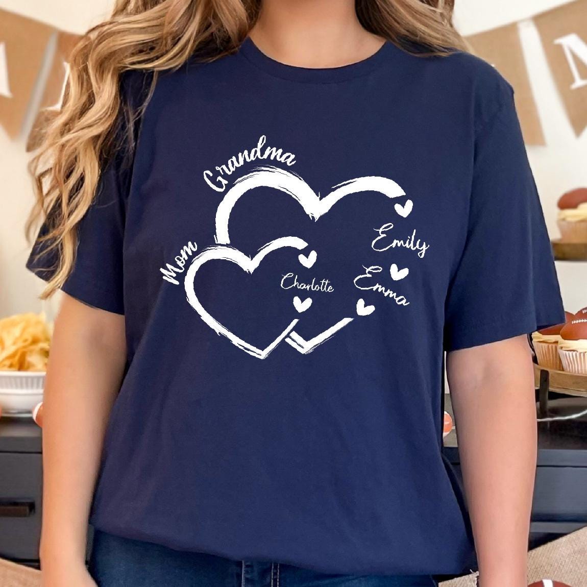 May include: Navy blue t-shirt with a white design of two interlocking hearts. The hearts are labeled with the words "Grandma", "Mom", "Emily", "Charlotte", and "Emma".