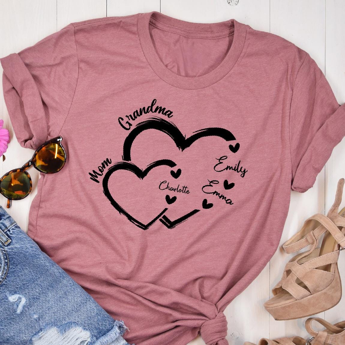 May include: A pink t-shirt with a design of two intertwined hearts. The hearts are outlined in black and have the words "Grandma", "Mom", "Emily", "Emma", and "Charlotte" written inside them.
