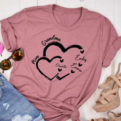 May include: A pink t-shirt with a design of two intertwined hearts. The hearts are outlined in black and have the words "Grandma", "Mom", "Emily", "Emma", and "Charlotte" written inside them.