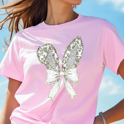 May include: A pink t-shirt with a white and gold bunny ear design. The ears are made of a floral pattern with a white bow in the center.