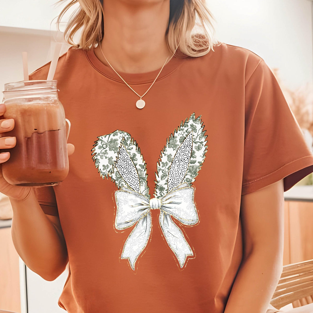 May include: A rust-colored t-shirt with a white bow and bunny ears design. The ears are decorated with a green and white floral pattern.