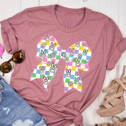May include: A pink t-shirt with a large pink bow printed on the front. The bow is made up of checkered squares in various pastel colors, with a pattern of smiling bunnies and flowers.
