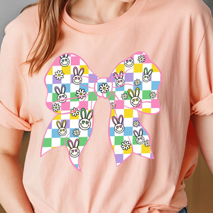 May include: A pink t-shirt with a large, colorful bow design. The bow is made up of checkered squares in pink, yellow, green, and purple, with bunny faces and flowers printed on them.