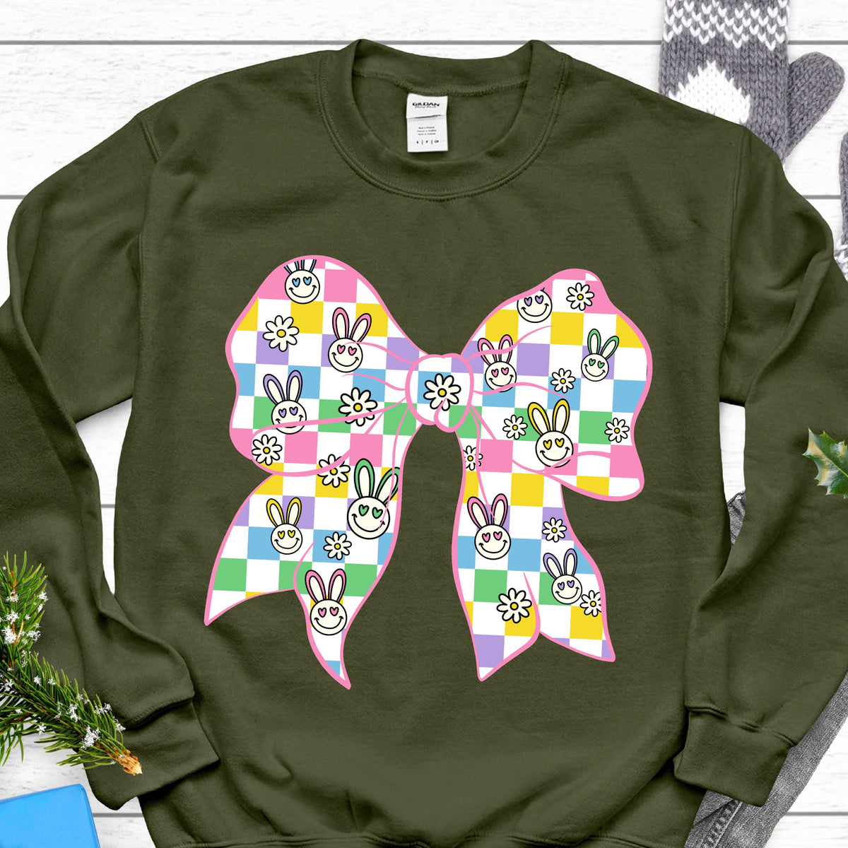 May include: A green sweatshirt with a pink and white checkered bow with smiling bunny faces. The bow has a green, yellow, and pink checkered pattern.