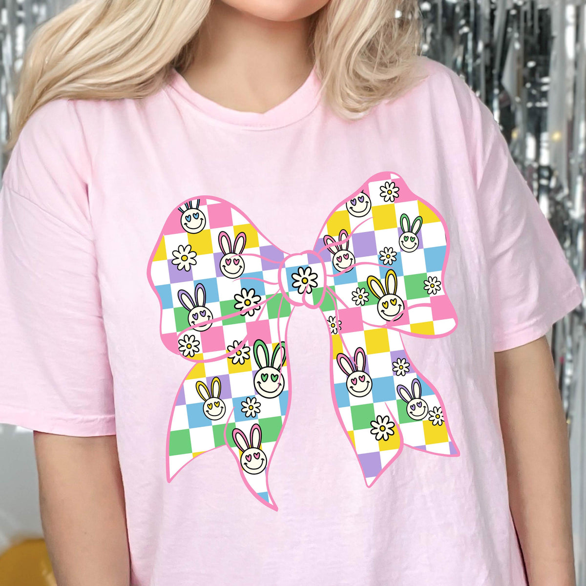 May include: A pink t-shirt with a colorful checkered bow featuring bunny faces and flowers. The bow is in a pastel color scheme with pink, yellow, green, blue, and purple.