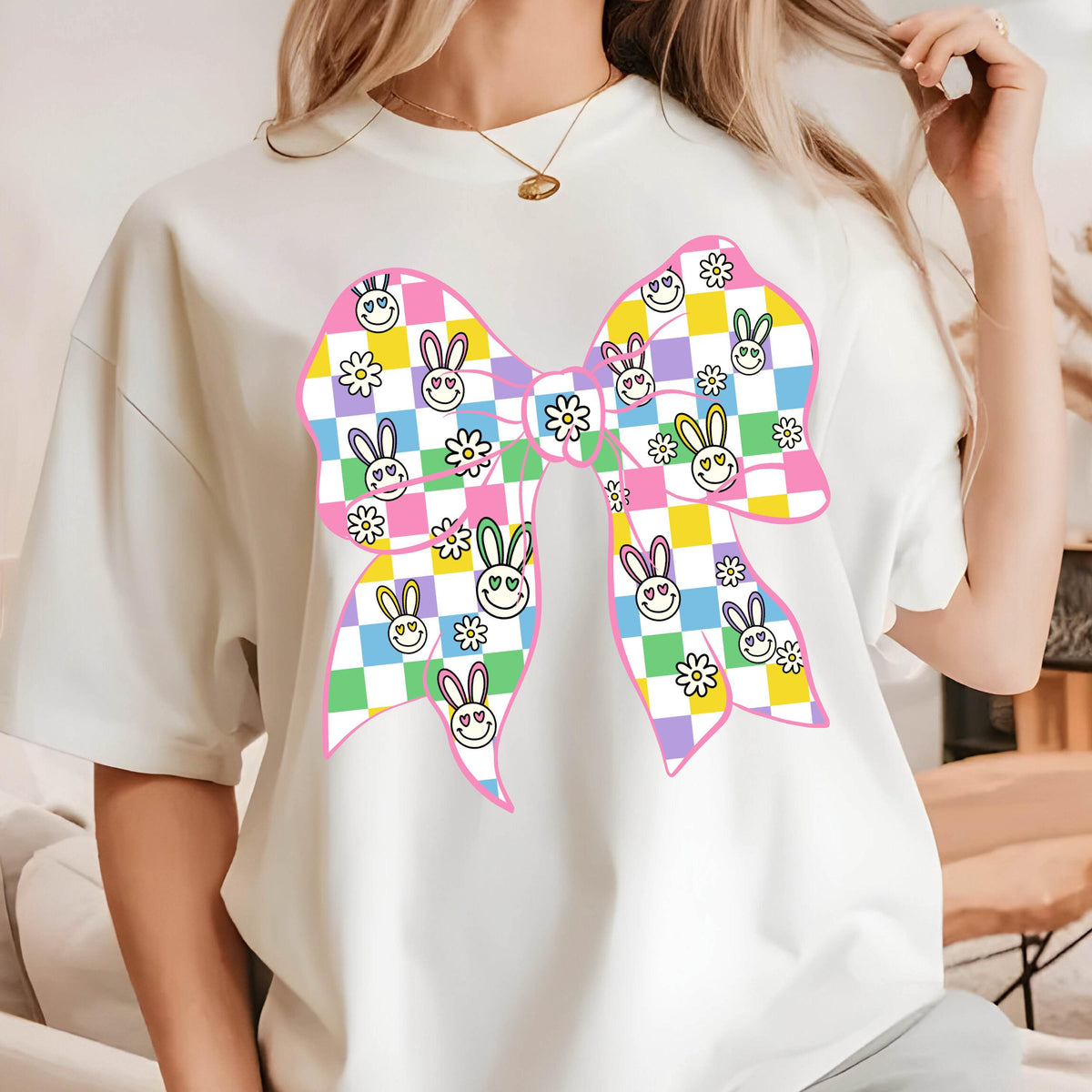 May include: White t-shirt with a pink bow design. The bow is made up of checkered squares in pink, yellow, green, and purple. Each square has a small bunny face with a smiley face.
