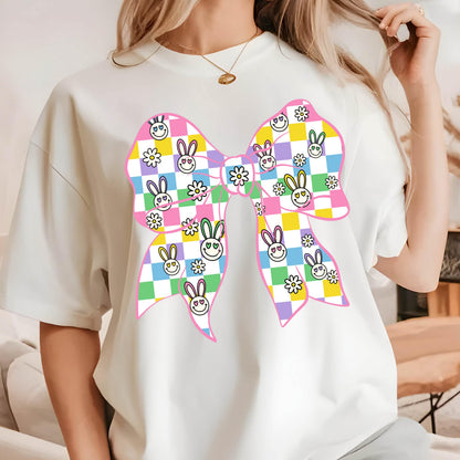 May include: White t-shirt with a pink bow design. The bow is made up of checkered squares in pink, yellow, green, and purple. Each square has a small bunny face with a smiley face.