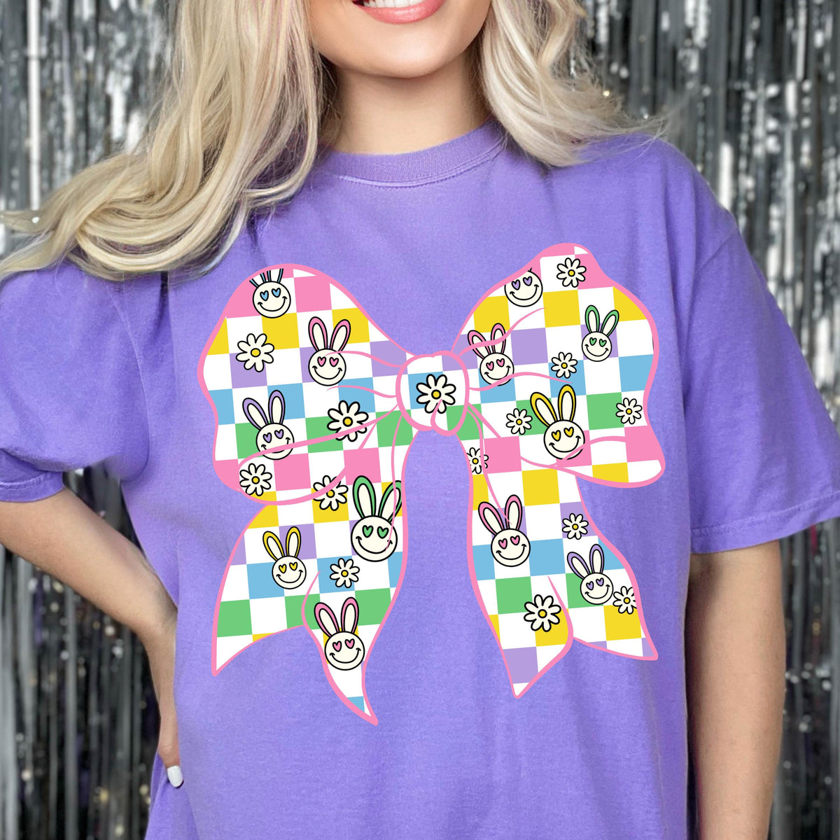 May include: Purple t-shirt with a pink bow design. The bow is covered in a checkered pattern of pastel colors with bunny faces and flowers.