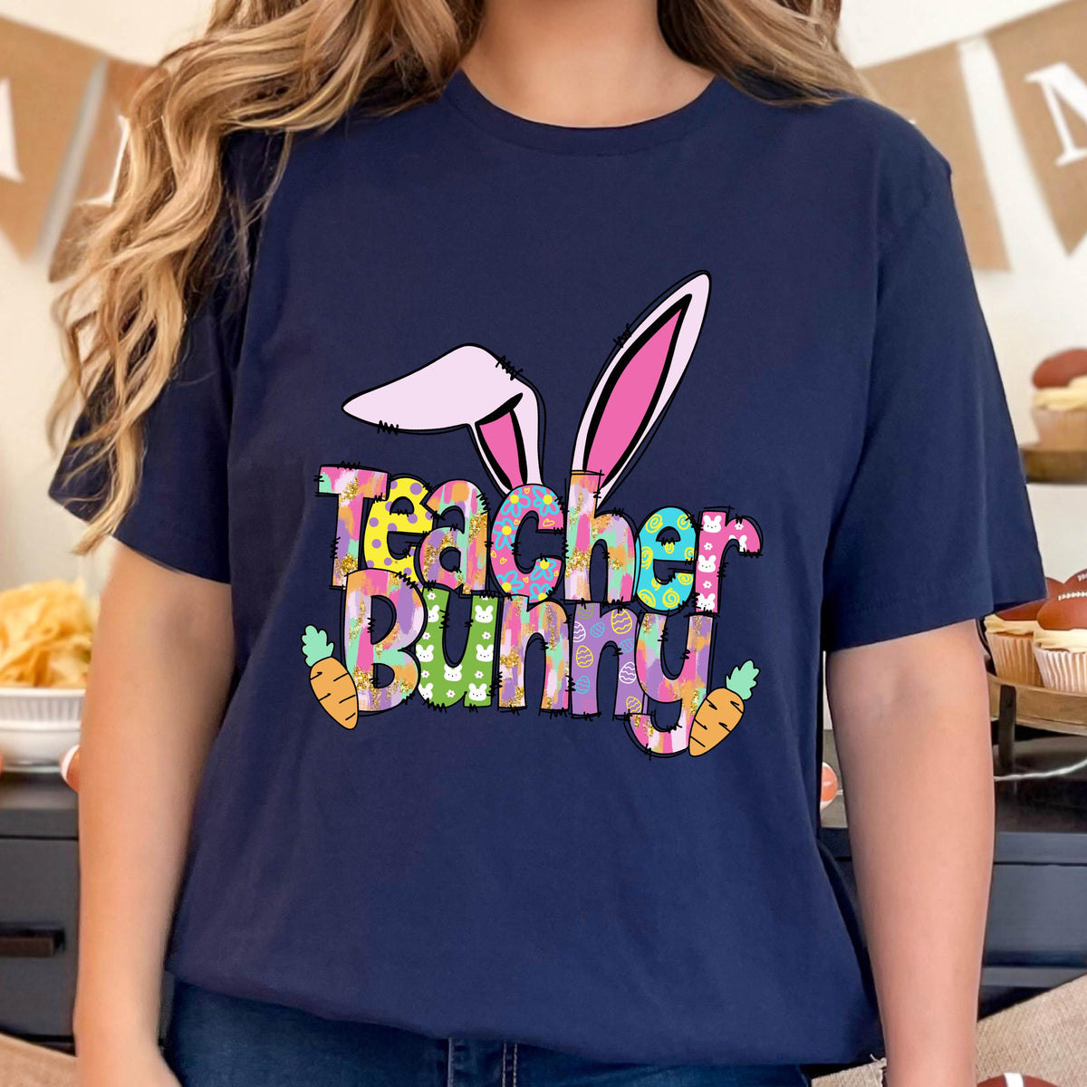 May include: Navy blue t-shirt with the words "Teacher Bunny" in colorful, patterned letters. The design includes bunny ears and two orange carrots, perfect for Easter or a teacher's gift.