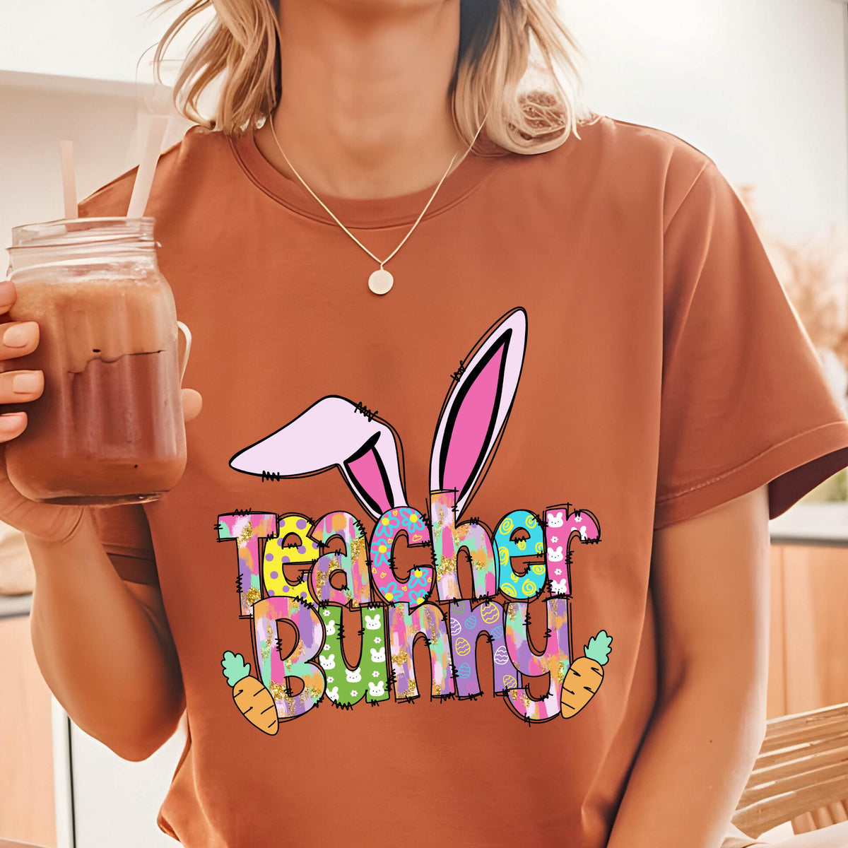 May include: A rust-colored t-shirt featuring a colorful graphic design. The design includes bunny ears, the words "Teacher Bunny", and carrot illustrations. The shirt is a casual style, suitable for Easter or teacher appreciation.