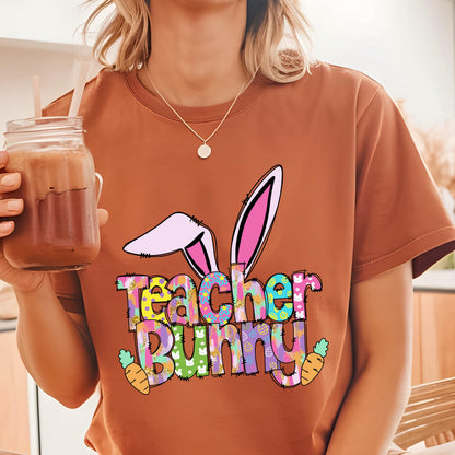 May include: A rust-colored t-shirt featuring a colorful graphic design. The design includes bunny ears, the words "Teacher Bunny", and carrot illustrations. The shirt is a casual style, suitable for Easter or teacher appreciation.