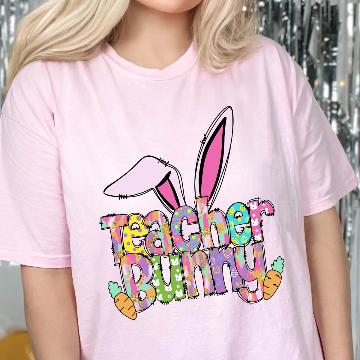 May include: A light pink t-shirt featuring a colorful graphic with the words "Teacher Bunny" and bunny ears. The design includes carrots and Easter egg patterns, perfect for teachers during the Easter season.