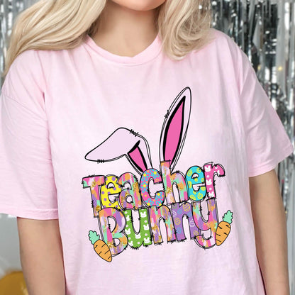 May include: A light pink t-shirt featuring a colorful graphic with the words "Teacher Bunny" and bunny ears. The design includes carrots and Easter egg patterns, perfect for teachers during the Easter season.
