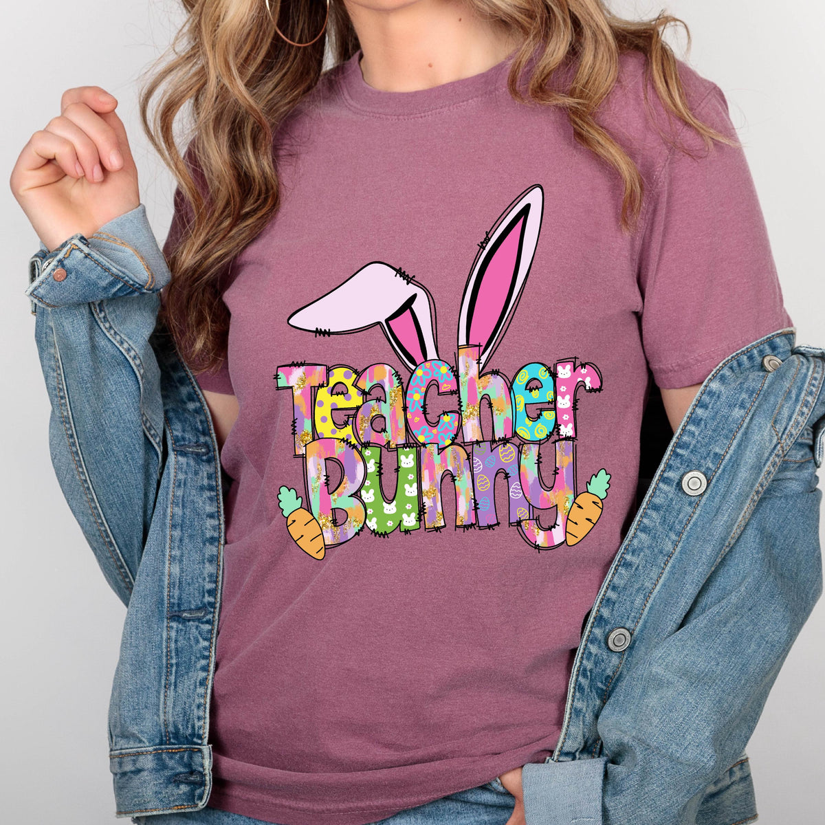 May include: A mauve-colored t-shirt with a colorful graphic that says "Teacher Bunny". The graphic includes bunny ears, carrots, and a mix of bright colors. The shirt is made of a soft material.