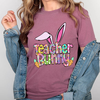 May include: A mauve-colored t-shirt with a colorful graphic that says "Teacher Bunny". The graphic includes bunny ears, carrots, and a mix of bright colors. The shirt is made of a soft material.