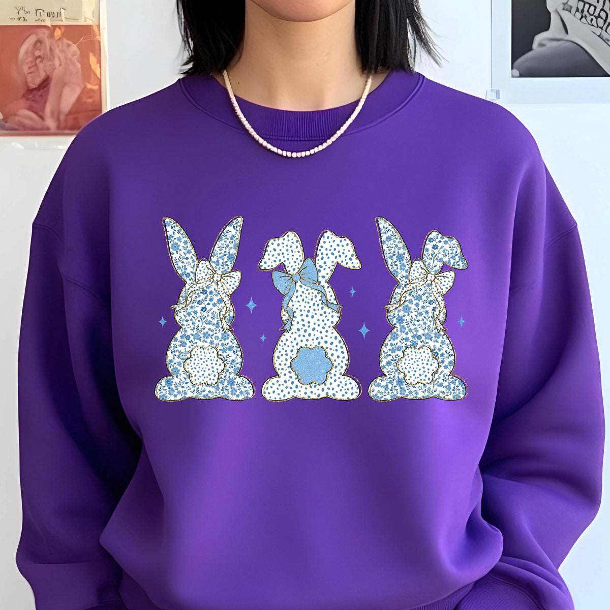 May include: Purple sweatshirt with a graphic of three blue and white bunny rabbits with bows. The bunnies are facing away from the viewer.