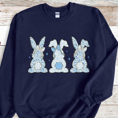 May include: A navy blue sweatshirt with a design of three blue and white bunny rabbits with bows. The bunnies are facing the viewer and are arranged in a row.