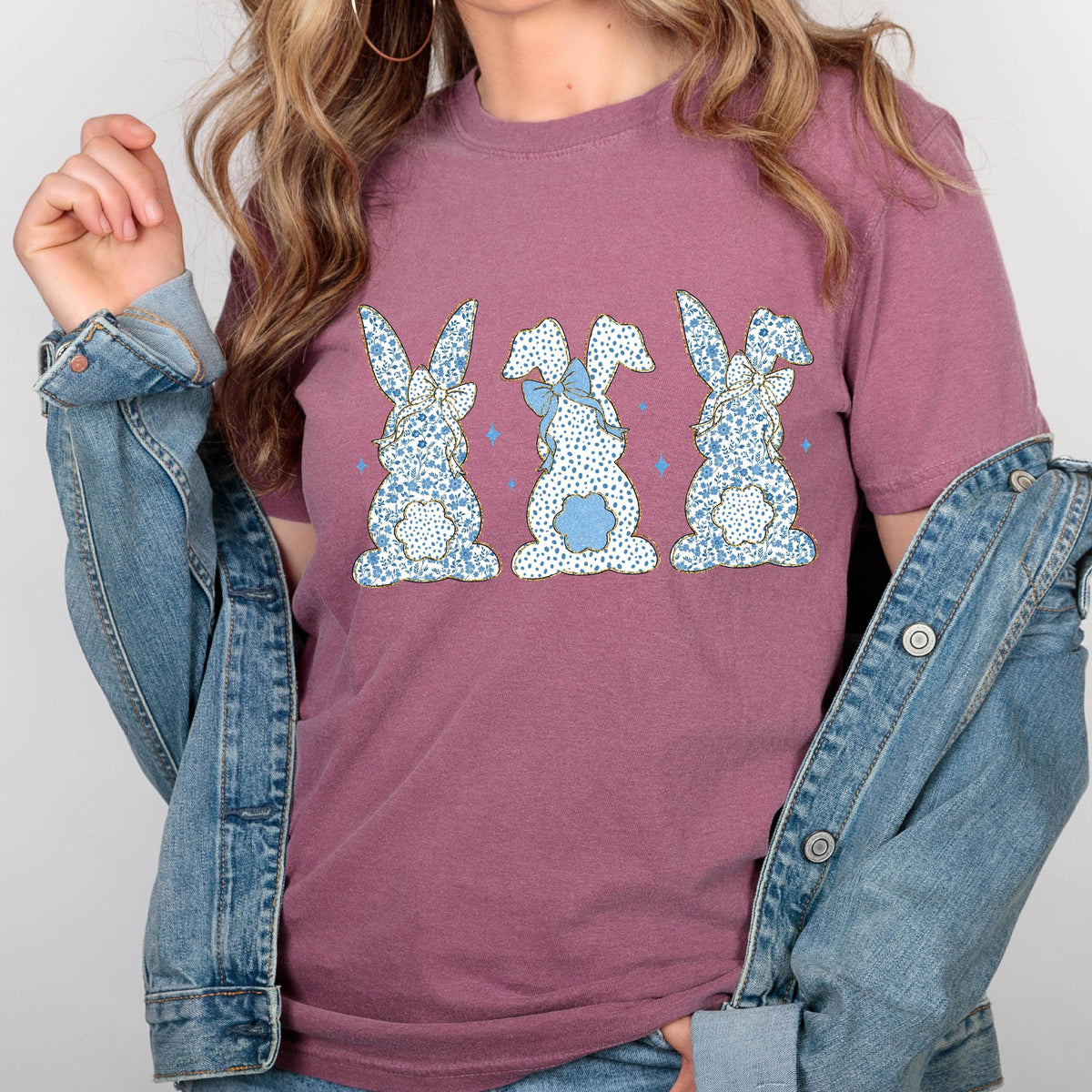 May include: A mauve t-shirt with a graphic of three blue and white patterned bunnies with blue bows. The bunnies are facing away from the viewer.