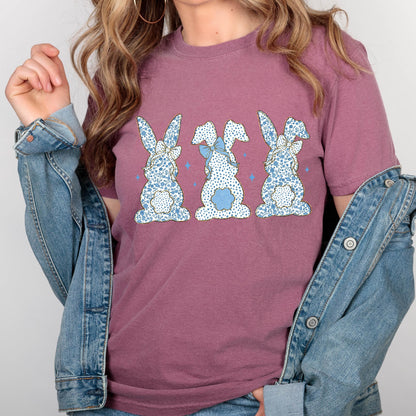 May include: A mauve t-shirt with a graphic of three blue and white patterned bunnies with blue bows. The bunnies are facing away from the viewer.