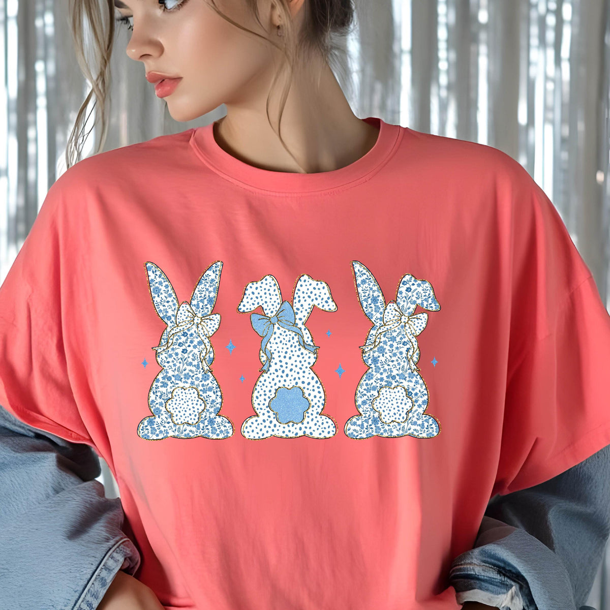 May include: A coral pink t-shirt with a graphic of three blue and white bunny rabbits with blue bows. The bunnies are facing away from the viewer.