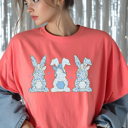 May include: A coral pink t-shirt with a graphic of three blue and white bunny rabbits with blue bows. The bunnies are facing away from the viewer.