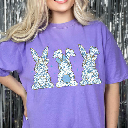 May include: Purple t-shirt with a graphic of three blue and white bunny rabbits with floral patterns and bows.