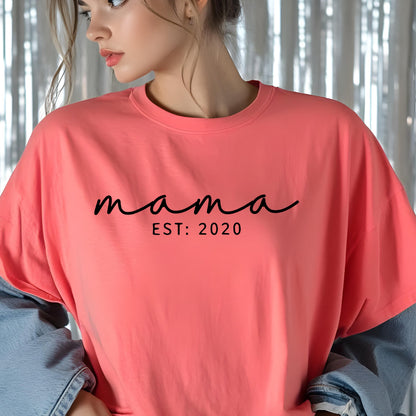 May include: A coral-colored t-shirt featuring the black text "mama" in a cursive script font, followed by the text "EST: 2020" in a simple sans-serif font centered on the chest. The garment has a crew neckline and short sleeves.