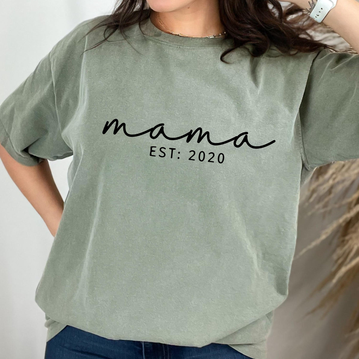 May include: Sage green t-shirt with the word "mama" in a cursive font and the text "EST: 2020" in a simple font. The shirt has short sleeves and a relaxed fit. The shirt is made of a soft, comfortable material.