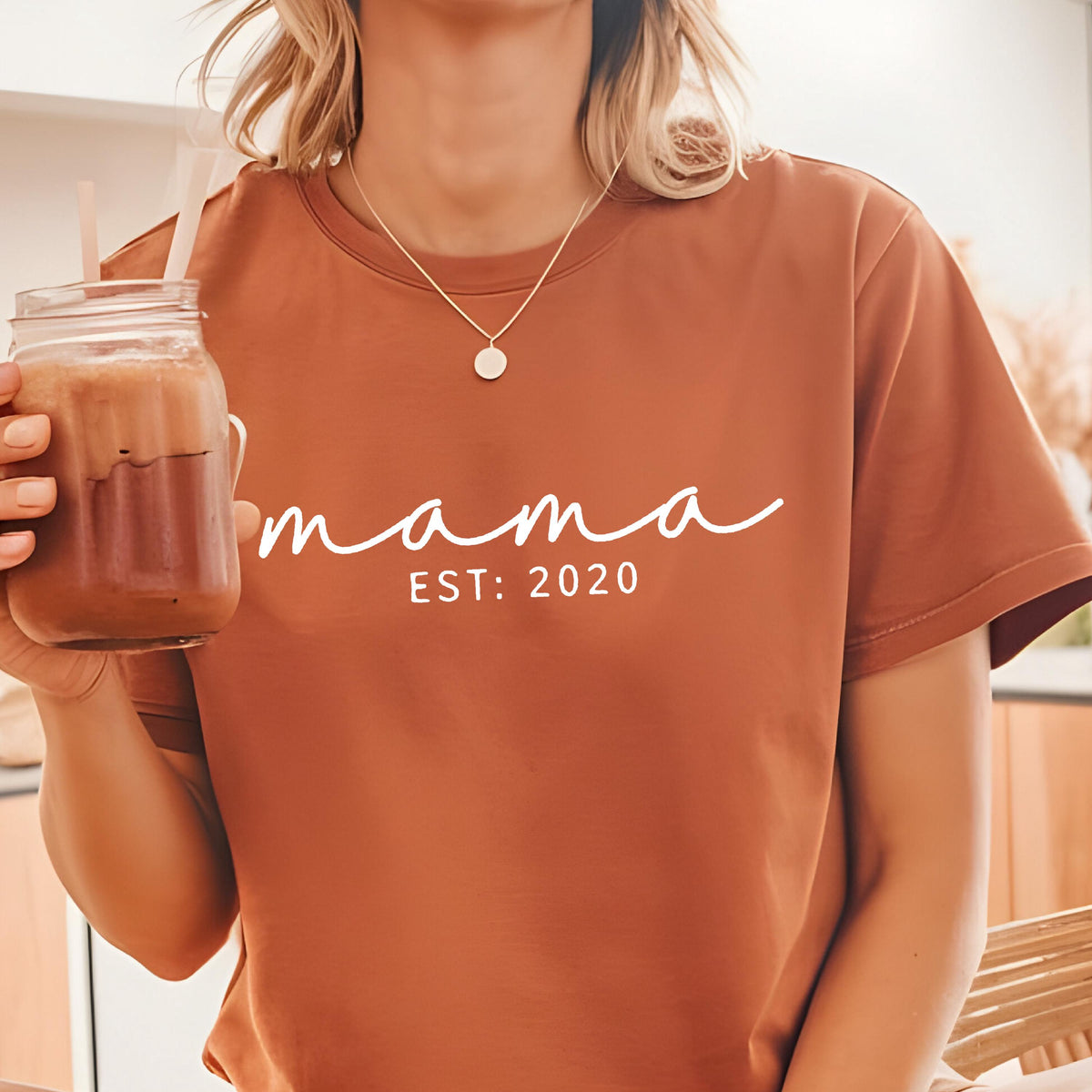 May include: A rust-colored t-shirt with the word "mama" in white script, and the text "EST: 2020" below it. The shirt has short sleeves and a round neckline. A person is holding a glass jar with a straw.