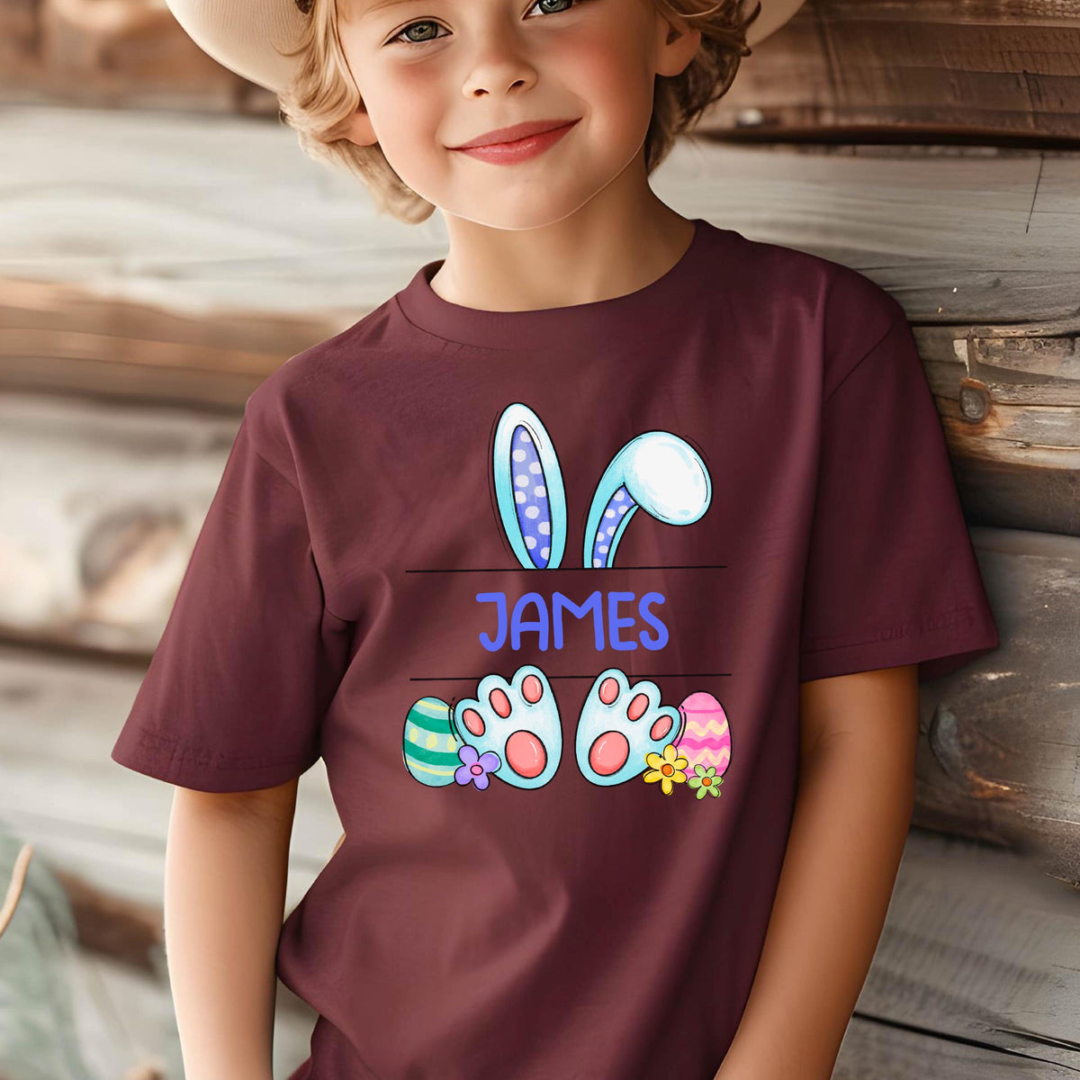 May include: A maroon short-sleeved t-shirt featuring a graphic of blue bunny ears, two bunny feet, and colorful Easter eggs. The name JAMES is printed in blue capital letters in the center between the ears and the feet of the bunny.