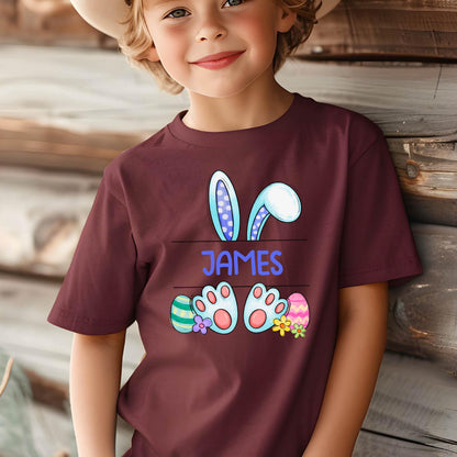 May include: A maroon short-sleeved t-shirt featuring a graphic of blue bunny ears, two bunny feet, and colorful Easter eggs. The name JAMES is printed in blue capital letters in the center between the ears and the feet of the bunny.