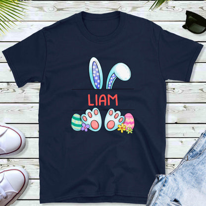 May include: A navy blue short-sleeve t-shirt featuring a graphic of light blue bunny ears, bunny feet, and colorful Easter eggs. The name LIAM is printed in red capital letters between the bunny ears and feet on the center of the shirt.