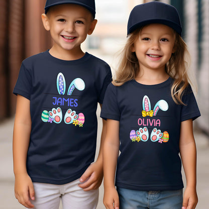 May include: Two navy blue t-shirts featuring a graphic of bunny ears, paws, and colorful Easter eggs. One shirt displays the name JAMES in blue text, while the other displays the name OLIVIA in pink text with a small yellow bow graphic.