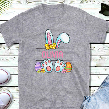 May include: A heather gray t-shirt featuring a graphic of blue and pink bunny ears with a yellow bow, the name OLIVIA in pink text, and a pair of blue bunny feet surrounded by colorful Easter eggs and small flowers on the lower section.
