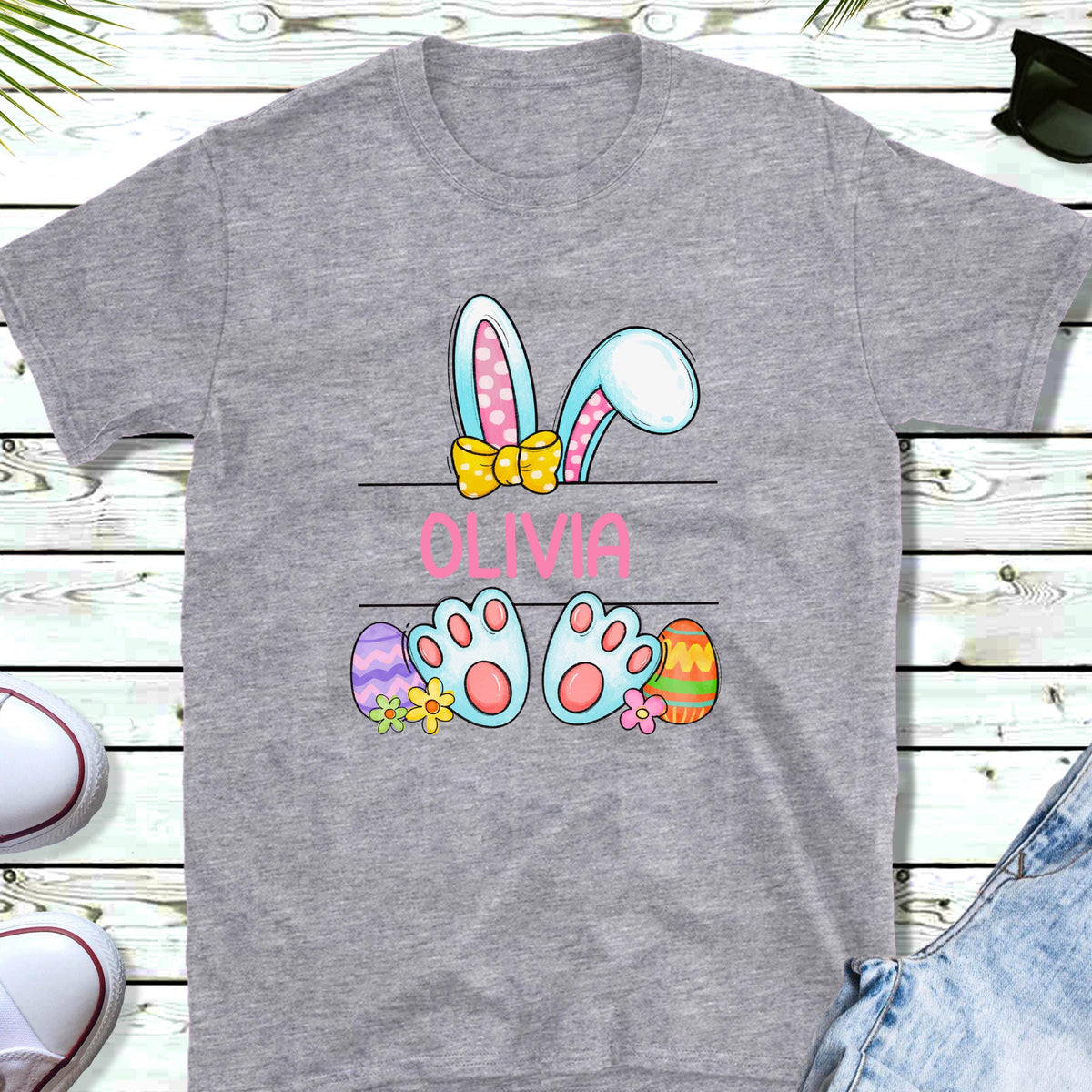 May include: A gray t-shirt with a cartoon bunny peeking over a sign that says "Olivia". The bunny has pink ears, a yellow bow, and blue feet. There are colorful Easter eggs on either side of the sign.