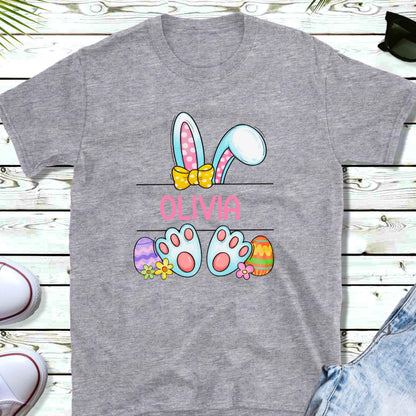 May include: A gray t-shirt with a cartoon bunny peeking over a sign that says "Olivia". The bunny has pink ears, a yellow bow, and blue feet. There are colorful Easter eggs on either side of the sign.