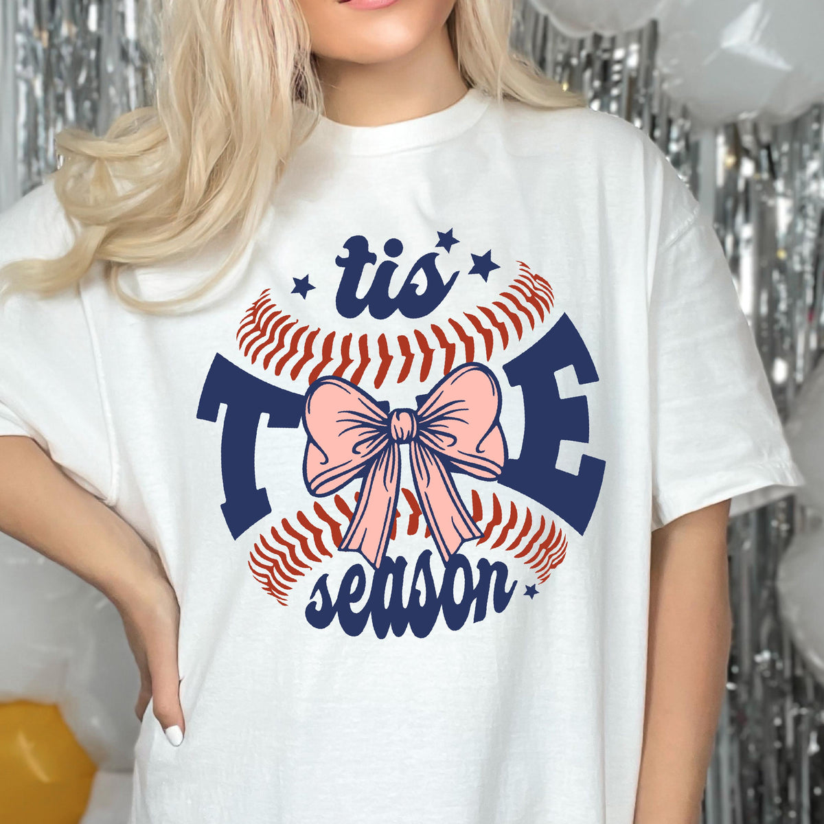 May include: White t-shirt with a baseball graphic and the text "tis the season" with a pink bow in the center.