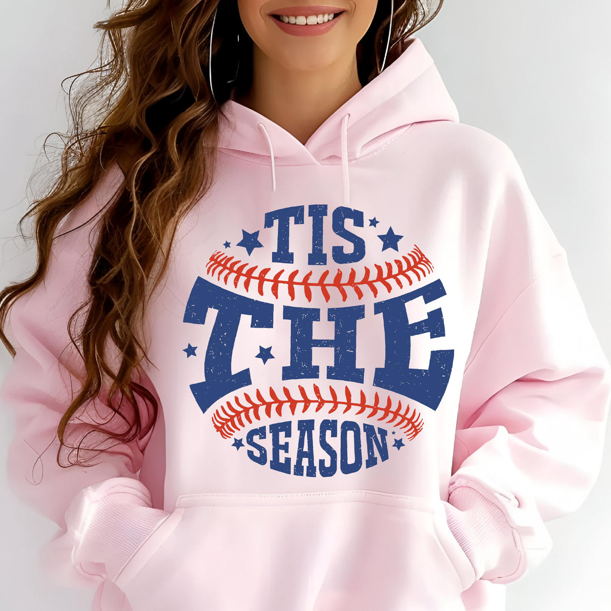 May include: A light pink hooded sweatshirt featuring a graphic of a baseball with red stitching and blue text that reads TIS THE SEASON. The design includes small blue stars and has a distressed texture. The hoodie has long sleeves and a front pocket.
