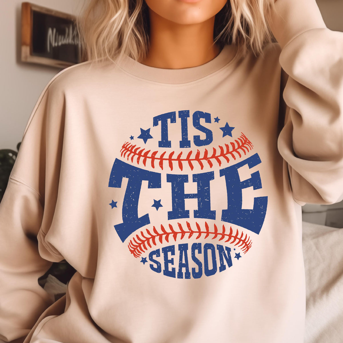 May include: A beige crewneck sweatshirt featuring a graphic of a baseball with red stitching and blue distressed text that reads TIS THE SEASON, accented with small blue stars arranged around the circular baseball design on the front.
