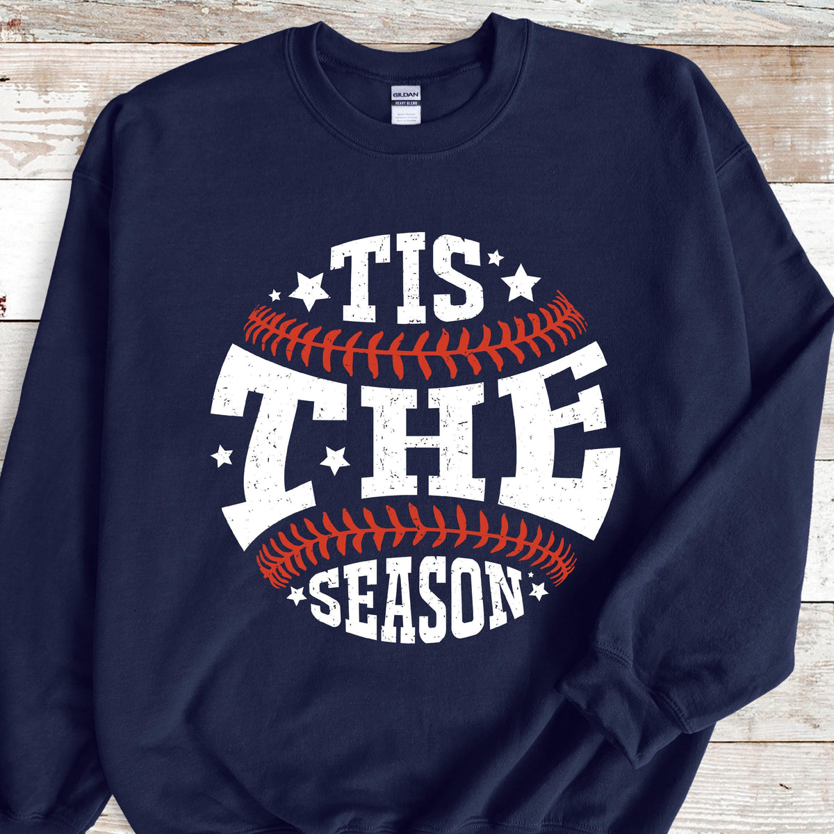 May include: A navy blue crewneck sweatshirt featuring a large graphic of a baseball with red stitching. The white distressed text reads TIS THE SEASON, arranged in a circular pattern around the baseball, accented by small white stars.