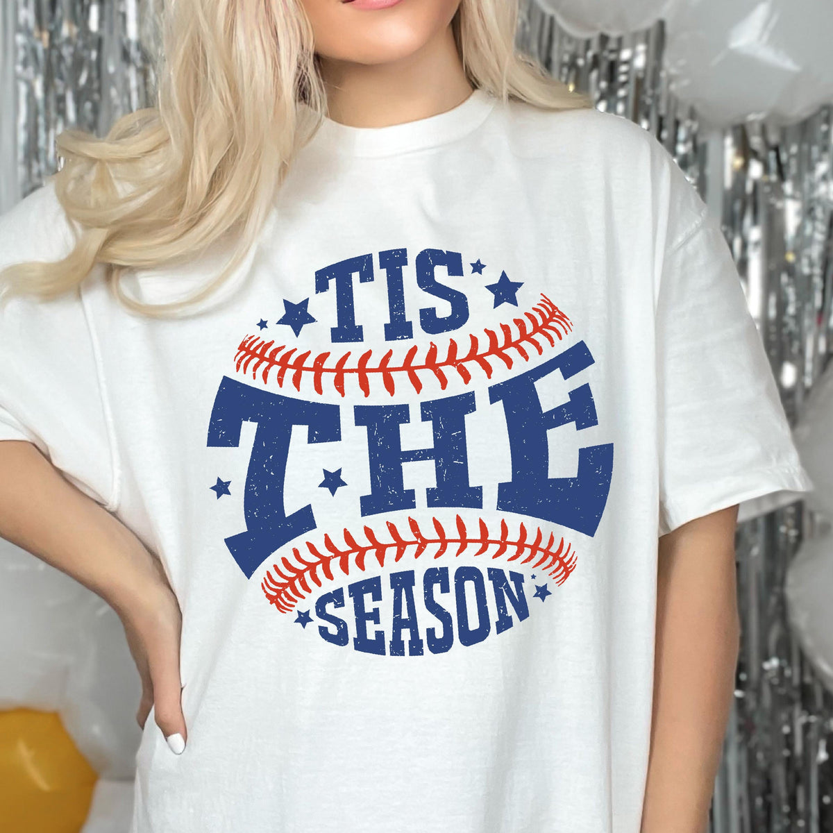 May include: White t-shirt with a blue and red baseball graphic and the text "Tis the Season" printed on it.