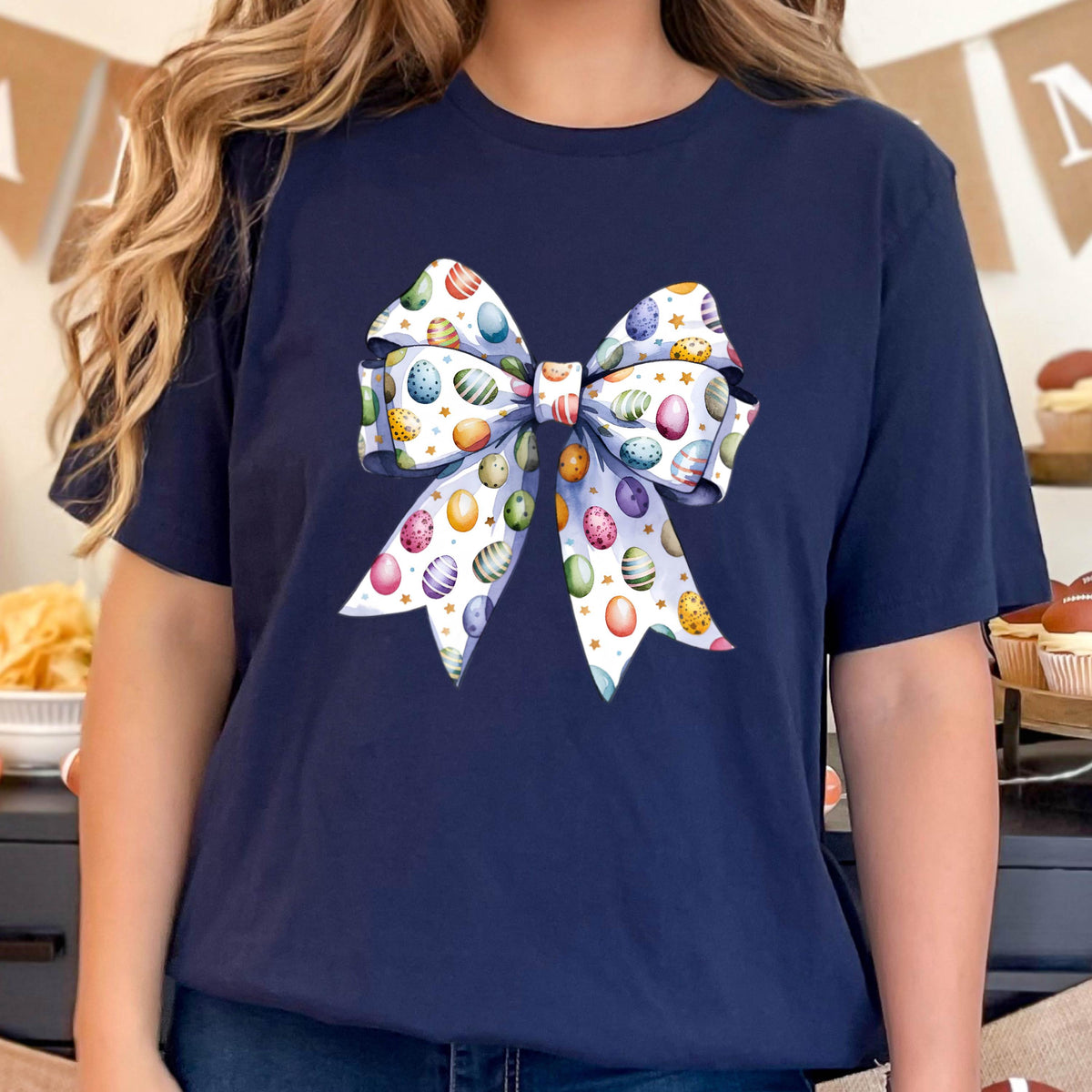 May include: A navy blue t-shirt with a white bow printed on the front. The bow is decorated with a pattern of colorful Easter eggs.