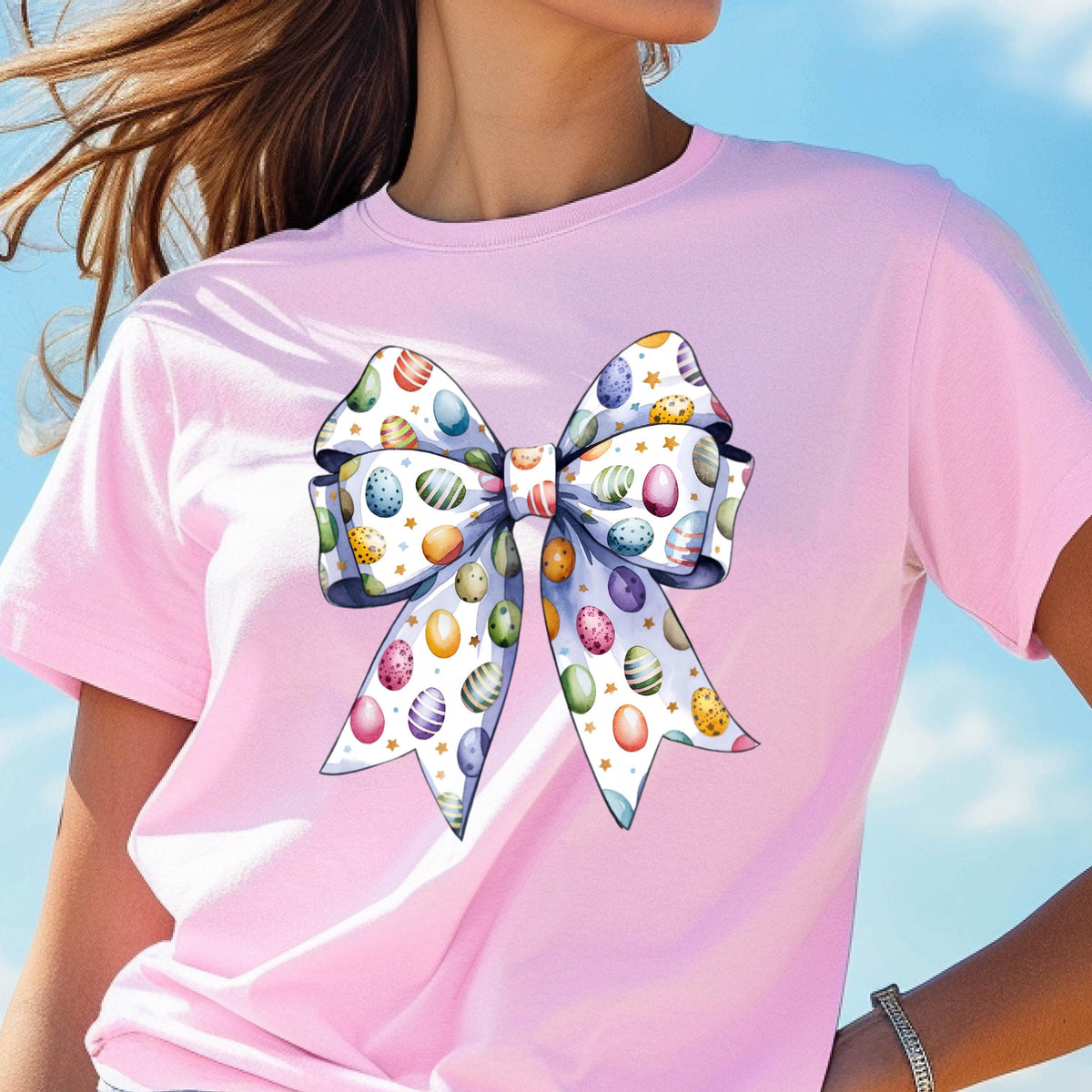 May include: A pink t-shirt with a white bow featuring a pattern of colorful Easter eggs. The bow is tied in a knot with the ends of the bow flowing down.