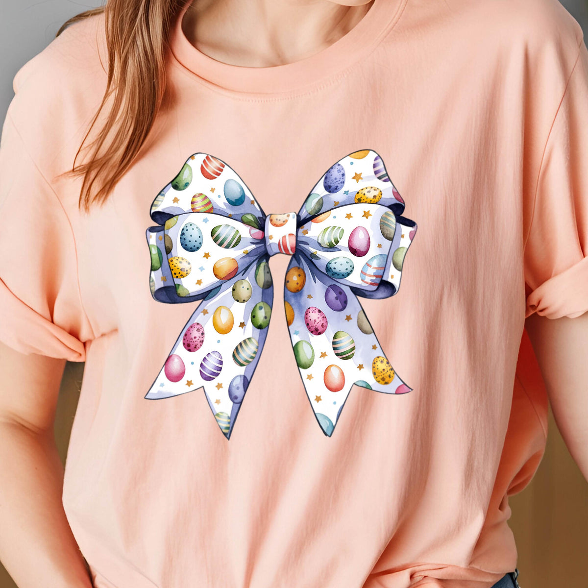 May include: A pink t-shirt with a large white bow decorated with colorful Easter eggs. The bow is tied in a knot and has a white background with gold stars.