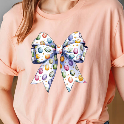 May include: A pink t-shirt with a large white bow decorated with colorful Easter eggs. The bow is tied in a knot and has a white background with gold stars.