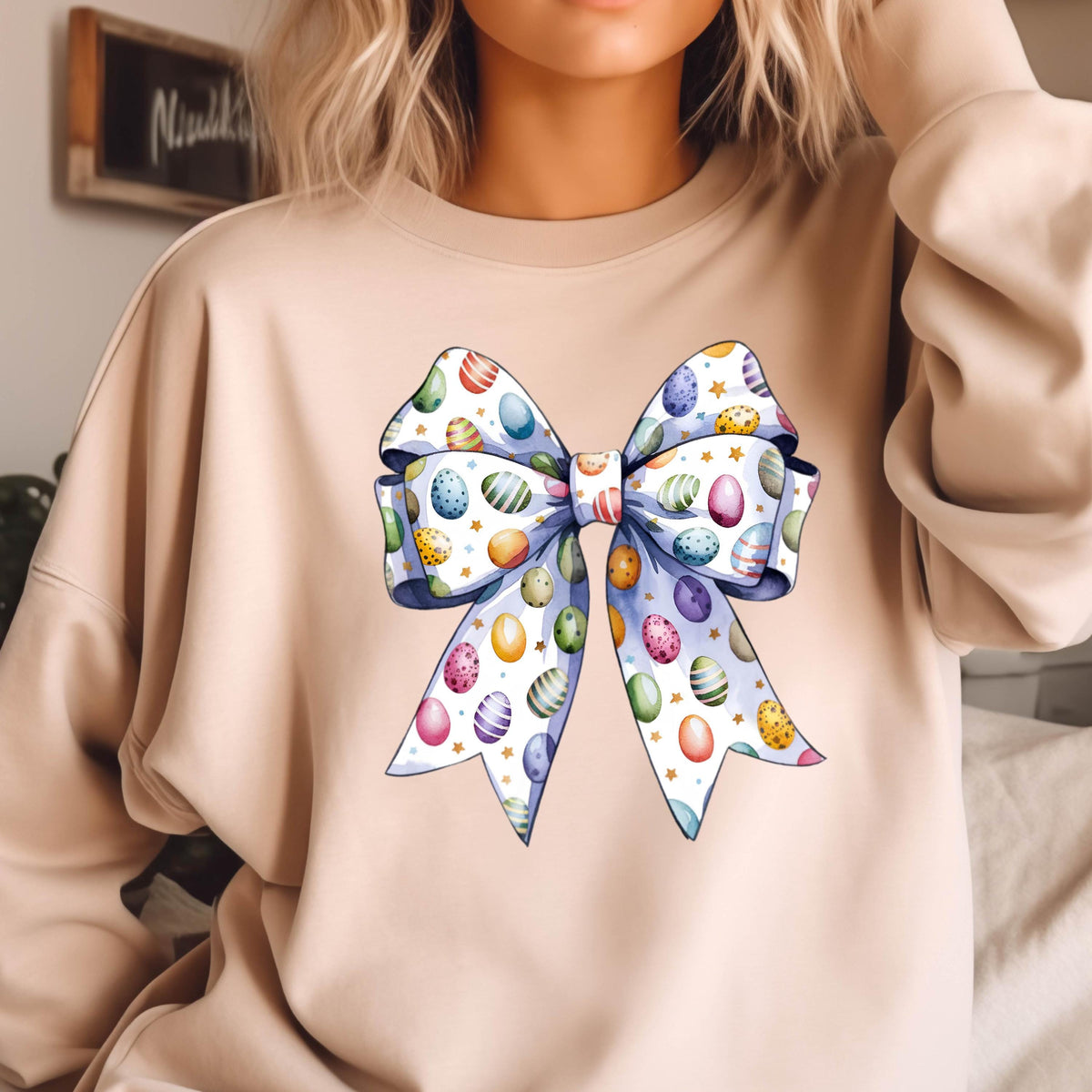 May include: A beige sweatshirt with a large blue bow printed on the front. The bow is decorated with colorful Easter eggs.