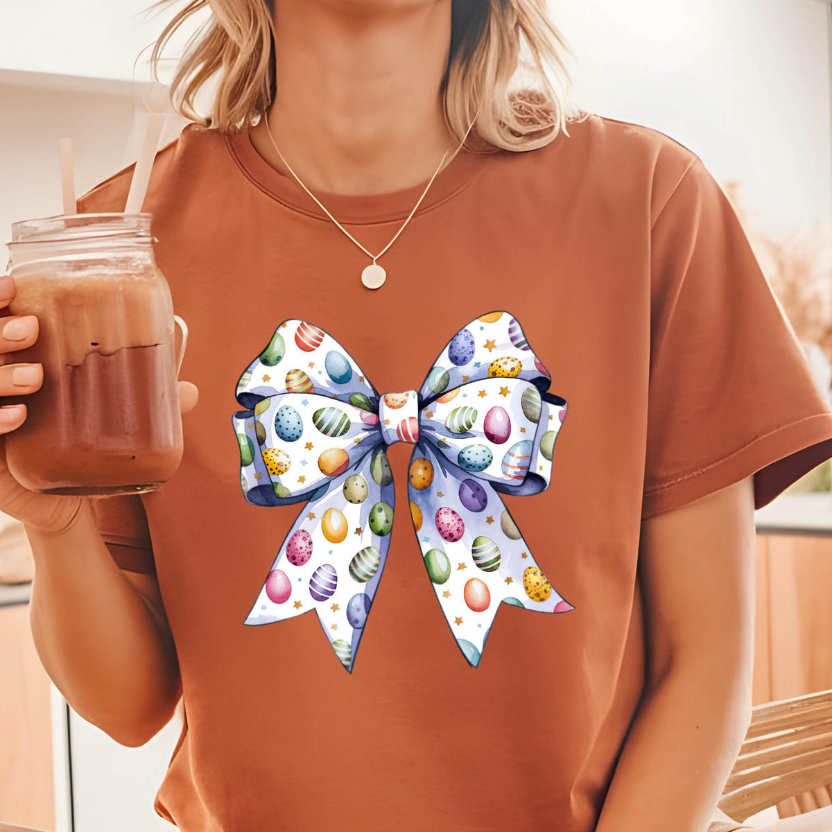 May include: A rust-colored t-shirt featuring a large, colorful bow design. The bow is decorated with Easter eggs in various colors and patterns, set against a light blue background. The shirt is short-sleeved and has a round neckline.
