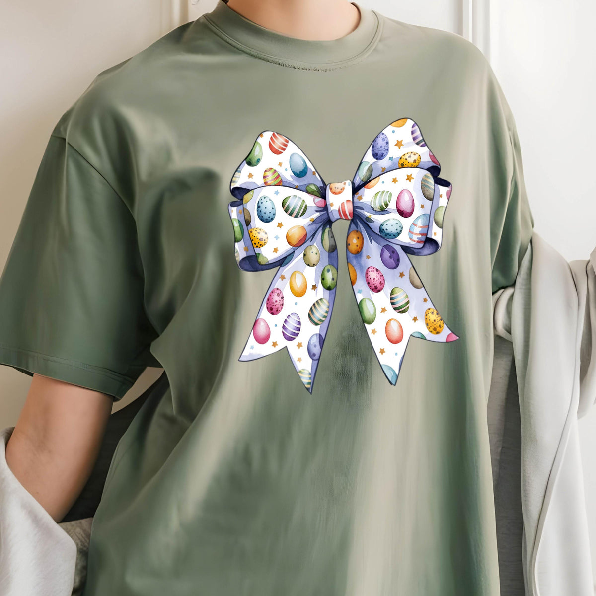 May include: Sage green t-shirt featuring a large bow design. The bow is decorated with colorful Easter eggs in various patterns and colors, including blue, purple, yellow, and pink. The bow has a white background with small gold stars.