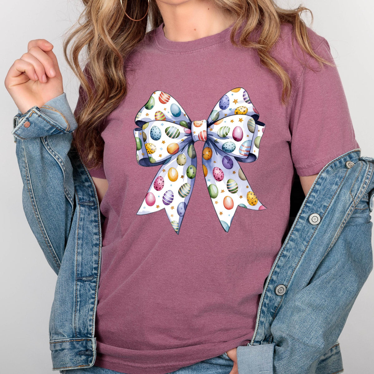 May include: A mauve-colored t-shirt featuring a large bow design. The bow is white with colorful Easter eggs and gold stars. The shirt is paired with a denim jacket.